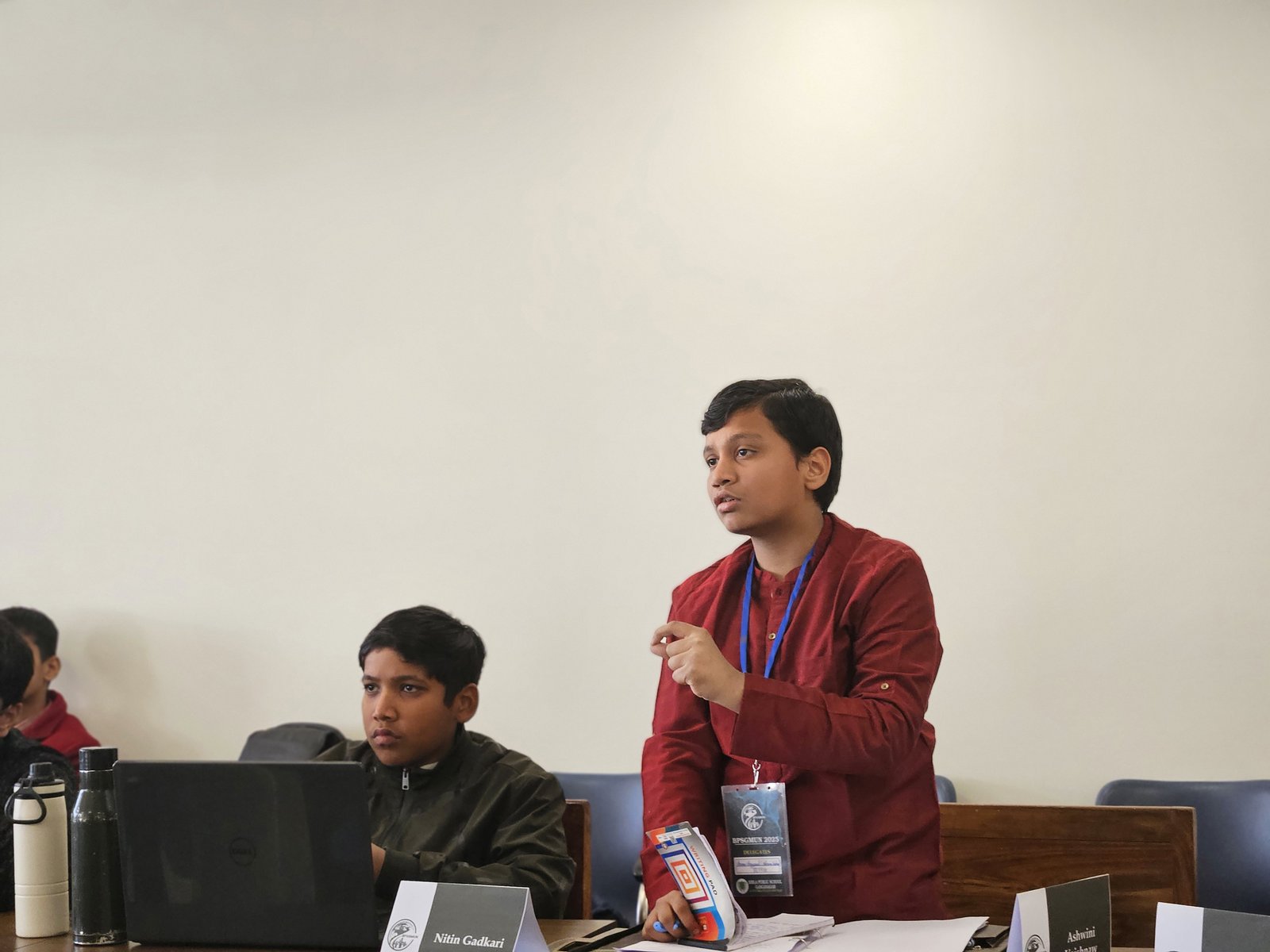 A delegate speaking, wearing a BPSGMUN 2025 delegate badge, with portfolio placards on the desk